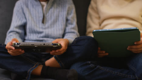 Nahaufnahme-Von-Zwei-Kleinen-Jungen,-Die-Zu-Hause-Auf-Dem-Sofa-Sitzen-Und-Spiele-Spielen-Oder-Auf-Ein-Digitales-Tablet-Oder-Ein-Tragbares-Spielgerät-Streamen